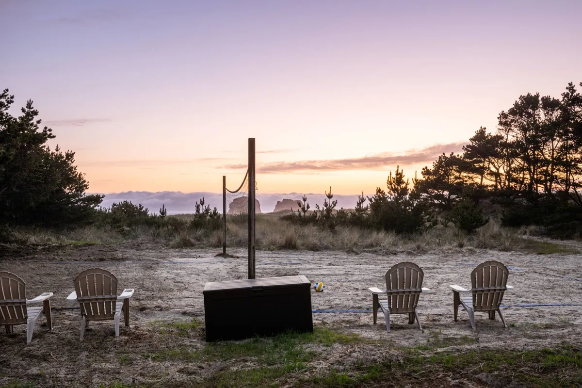 Adirondack chairs are arranged beside a sandy volleyball court with a net, all set against a stunning backdrop featuring ocean views and distant cliffs. The mood feels peaceful but social, inviting both relaxation and active play as the sun sets over the waves.