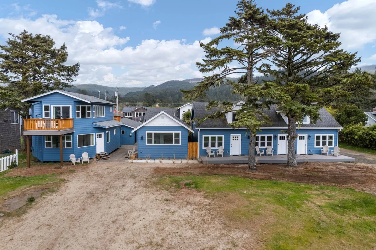 A welcoming exterior view of a blue house featuring a deck with chairs, large trees, and an open yard area. The space looks ready for family gatherings, outdoor relaxation, and includes references to stories around a fire pit.