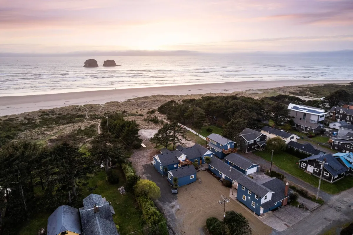 This image captures a sweeping view of the ocean with the iconic Twin Rocks arch prominently visible in the distance. The sandy expanse of Rockaway Beach stretches out, bordered by a cluster of houses, conveying the property's beachfront location and proximity to the natural landmark.