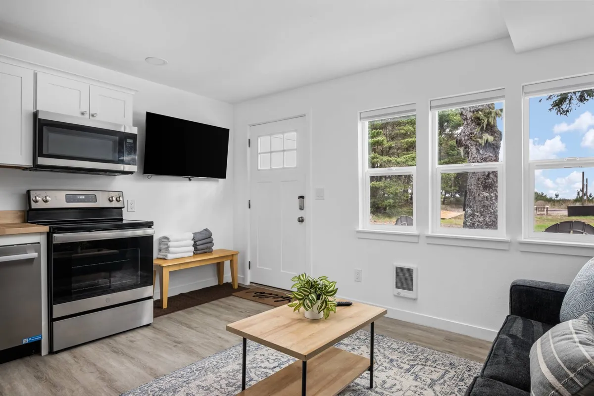A wide view of the open-concept living and kitchen area, featuring a stylish couch, modern kitchen appliances, a dining table, and large windows that fill the space with natural light. The décor is contemporary, and the furnishings look new and inviting, creating a seamless flow ideal for both relaxing and entertaining.