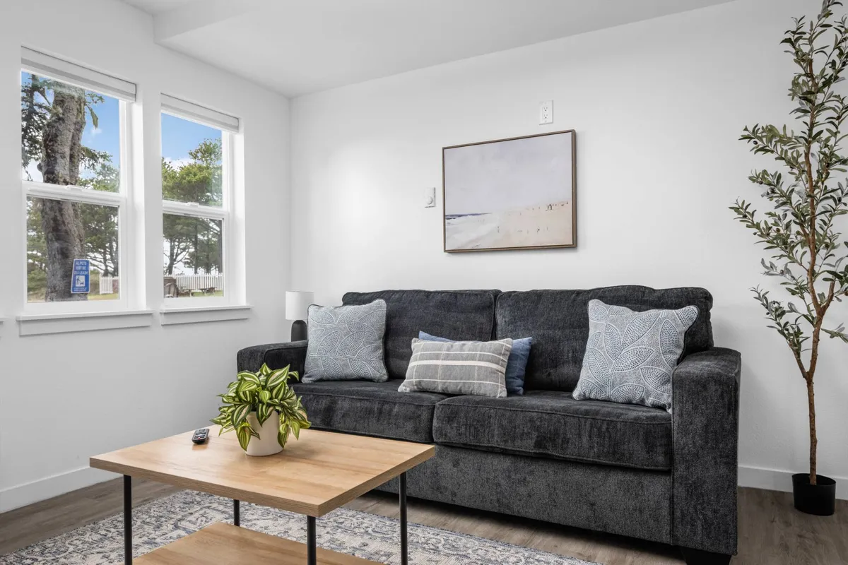 A cozy corner of the living room with a plush sofa facing a coffee table, artwork on the walls, and large windows that offer glimpses of the outdoors. The mood is warm and inviting, ideal for lounging, reading, or enjoying the coastal view through the window.