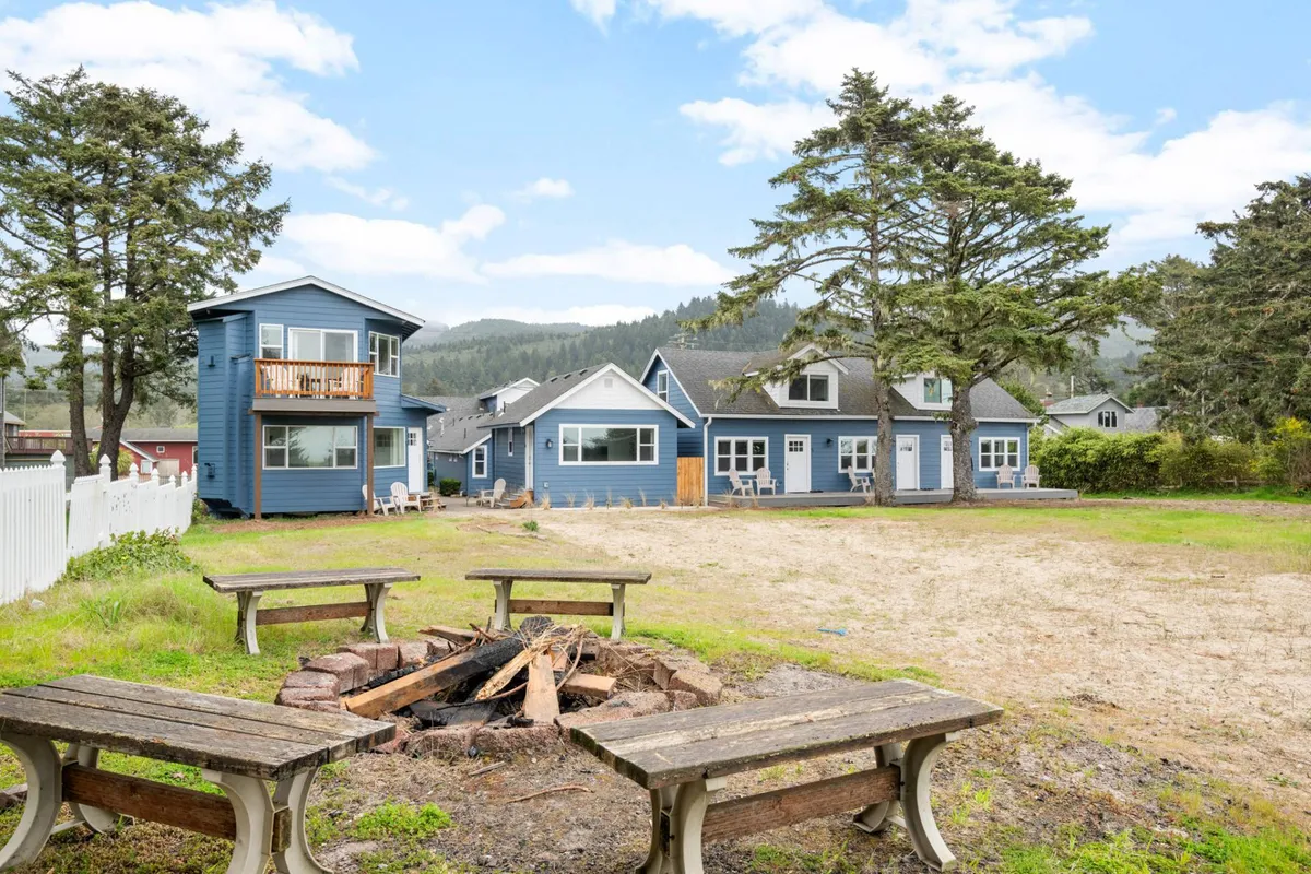 A cozy fire pit area is surrounded by blue houses and tall trees. Adirondack chairs are grouped around the fire pit, creating an inviting space for guests to gather, share stories, and make s’mores as the sun sets behind the mountains.
