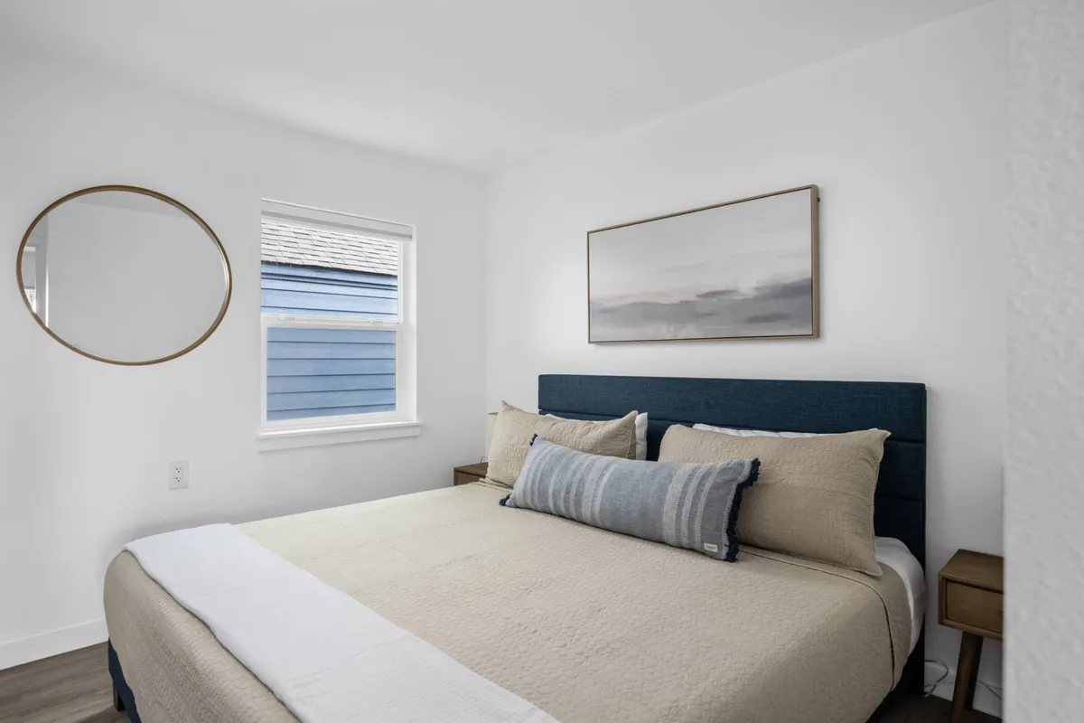 A serene and cozy bedroom scene with a plush bed, decorative mirror, and calming artwork above the headboard. Natural light fills the space, amplifying the sense of tranquility and privacy.