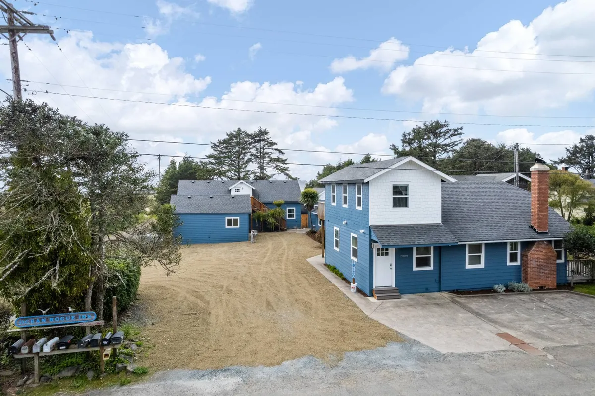 A wide-angle view of blue houses, a gravel driveway, and a prominent signpost, with space for outdoor gatherings. The setting is open and suggests a connection to a broader community and nearby nature, ideal for groups and family activities.