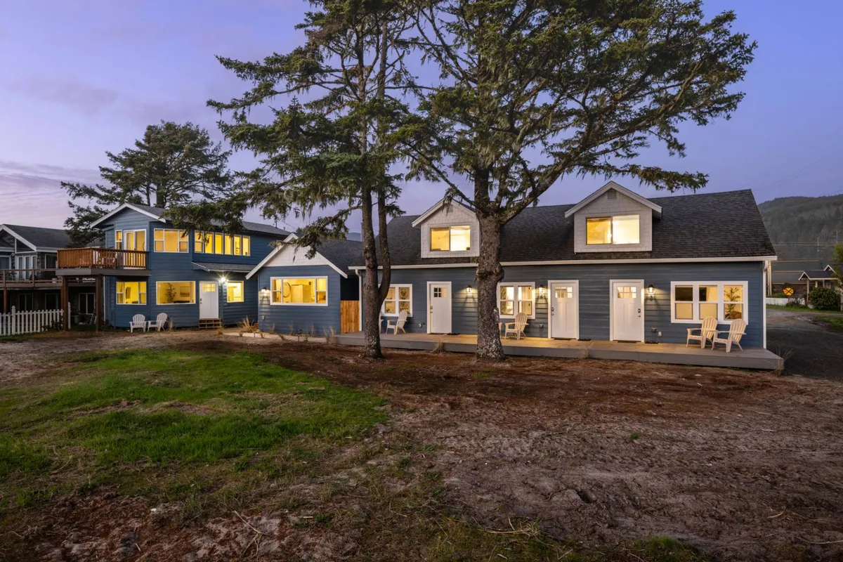 This image captures the blue exterior of the house with warmly illuminated windows, surrounded by lush trees and a welcoming front porch. The scene evokes a sense of arrival and comfort, showing the home as a cozy retreat after a day of exploring downtown Rockaway Beach.