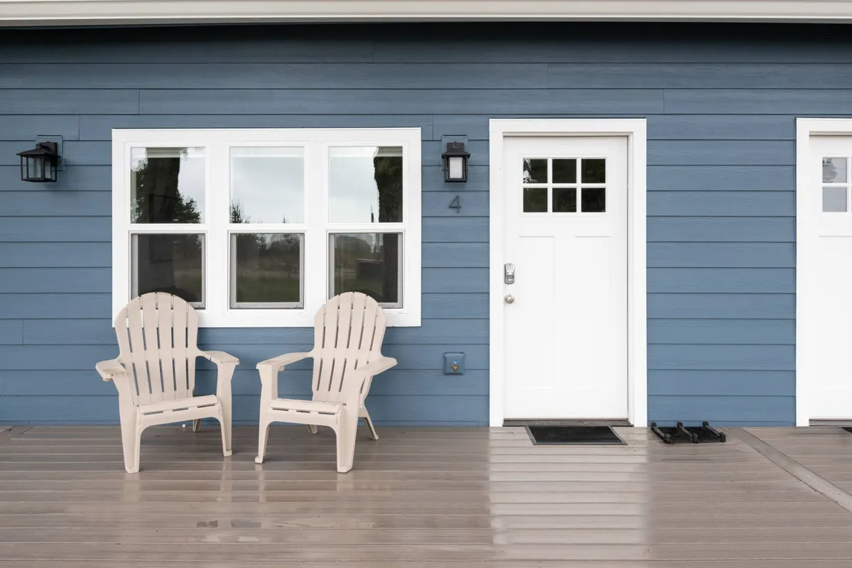 The inviting exterior features two beige Adirondack chairs on a small porch by the house’s white front door and blue wooden siding. The space is bathed in natural light, with clear access to grassy outdoor areas just steps away.